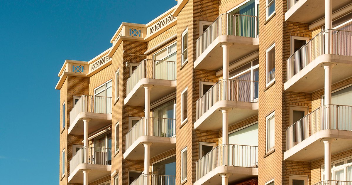 Tan brick mid-rise condominium building with rows of white balconies against a blue sky