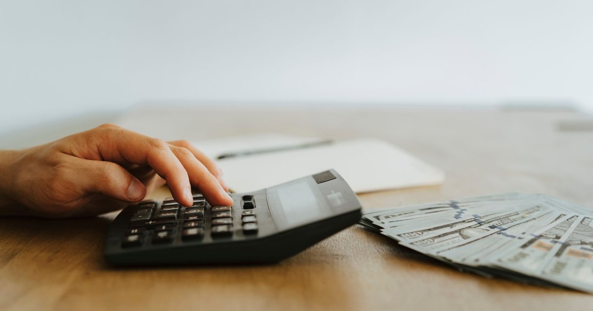 Hand pressing keys on a black desktop calculator beside a fan of dollar bills
