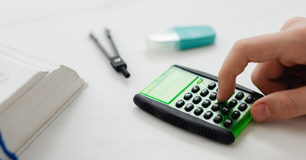 Hand pressing keys on a green desktop calculator on a white desk beside a notebook